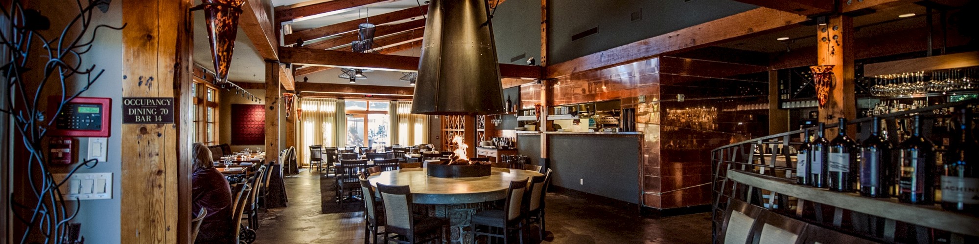 The image shows a modern restaurant interior with wooden beams, tables, chairs, and a bar area, creating a cozy dining atmosphere.