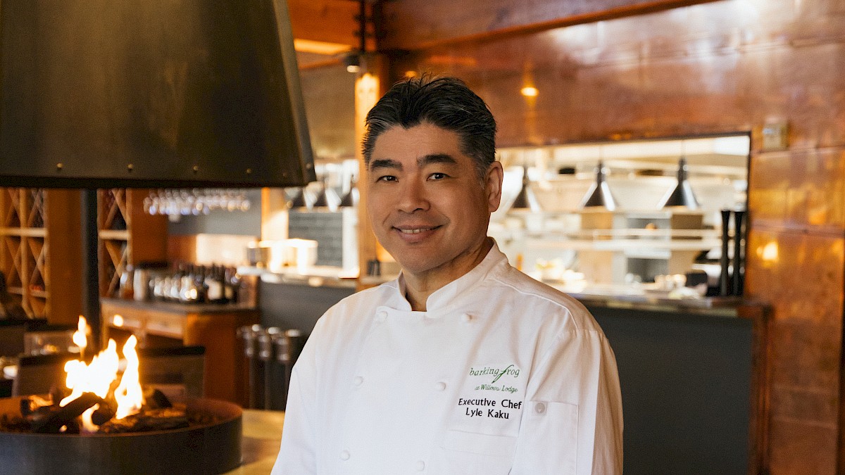 A chef in a white coat stands in a restaurant with a modern interior and a warm, open fire feature in the background.